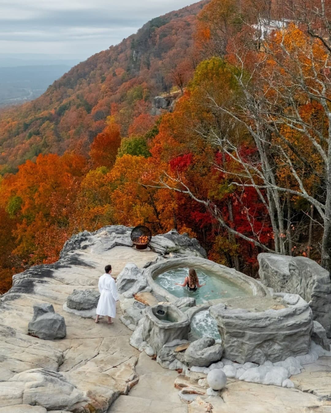 Couple at cliff-edge stone hot tub surrounded by vibrant fall foliage at BOLT FARM Treehouse