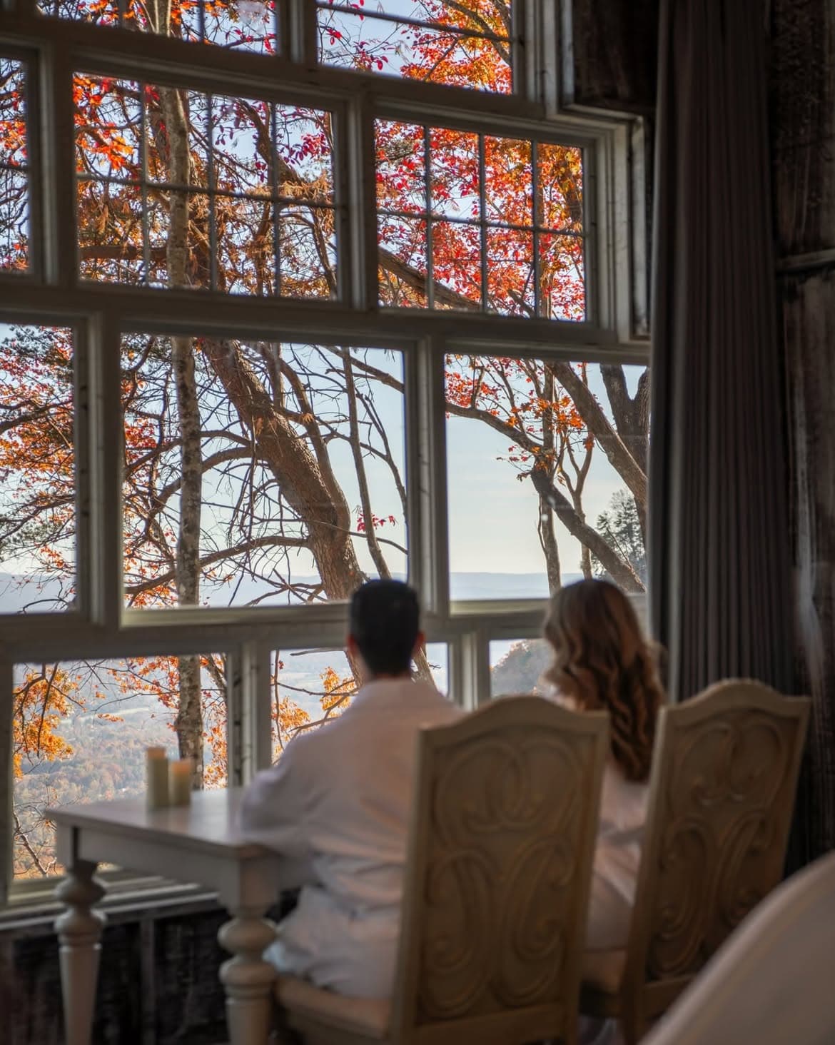 Couple in robes gazing out at fall foliage through treehouse windows